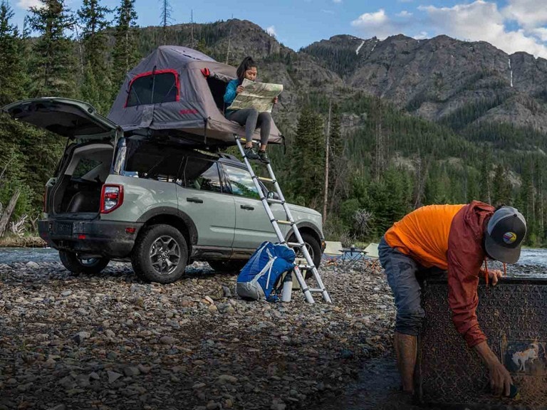 A grey Ford Bronco® Sport parked on a riverbed with tent on top as a couple plans a hike.