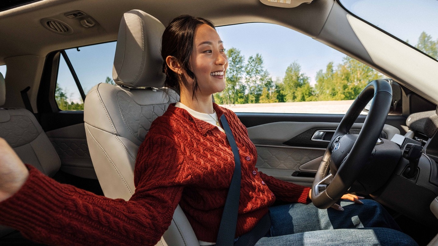A woman smiles while sitting in the driver’s seat of a Ford Explorer with her hands relaxed and off the steering wheel, utilizing hands-free driving. She is wearing a red knit sweater and jeans, and appears calm and content. The car interior features light-colored leather seats with a geometric pattern, and the background shows a clear, sunny day with trees visible through the windows.