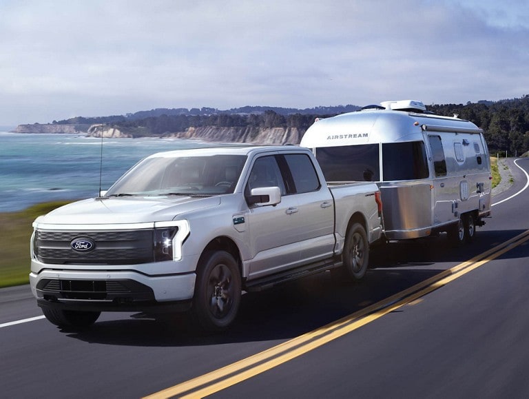 A silver Ford F-150® Lightning® truck being driven on a seaside highway while towing a silver trailer.