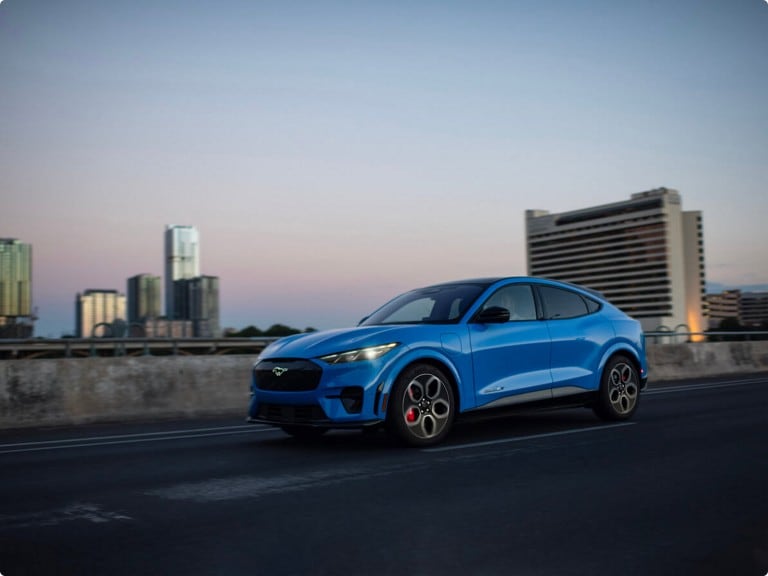 A blue Ford Mustang® Mach-E® SUV being driven on a city highway at dusk.