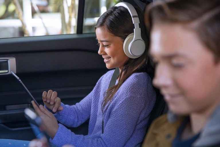 Two children with seatbelts in the back seat of a Ford vehicle looking at their mobile devices and listening to headphones.