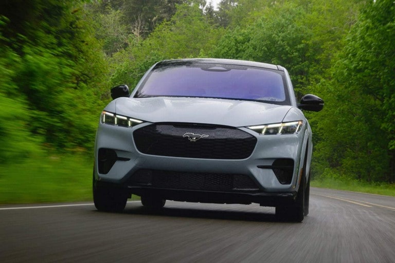 A blue Ford Mustang Mach-E® SUV being driven on a tree-lined country road surrounded by greenery.