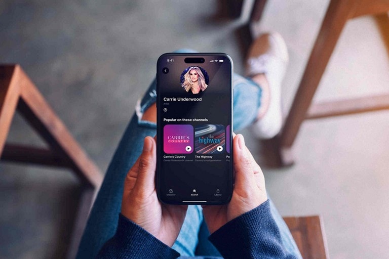 A listener holding a mobile phone device showing a SiriusXM mobile app screen.