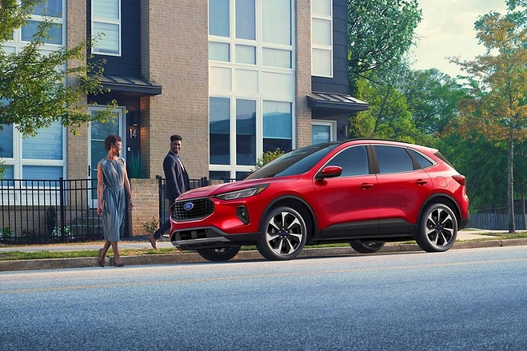 A man and woman walking up to a red Ford SUV parked on the side of a street