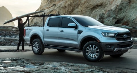 Un hombre colocando una tabla de surf en el portaequipajes de una Ford Ranger LARIAT 2020 estacionada sobre la arena cerca de una playa rocosa