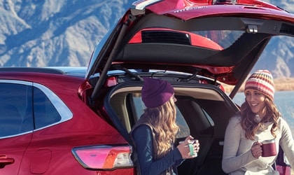 Dos mujeres sentadas en la parte trasera de una SUV Ford Escape® 2022 estacionada frente a un espejo de agua tomando café mientras un hombre cercano está pescando