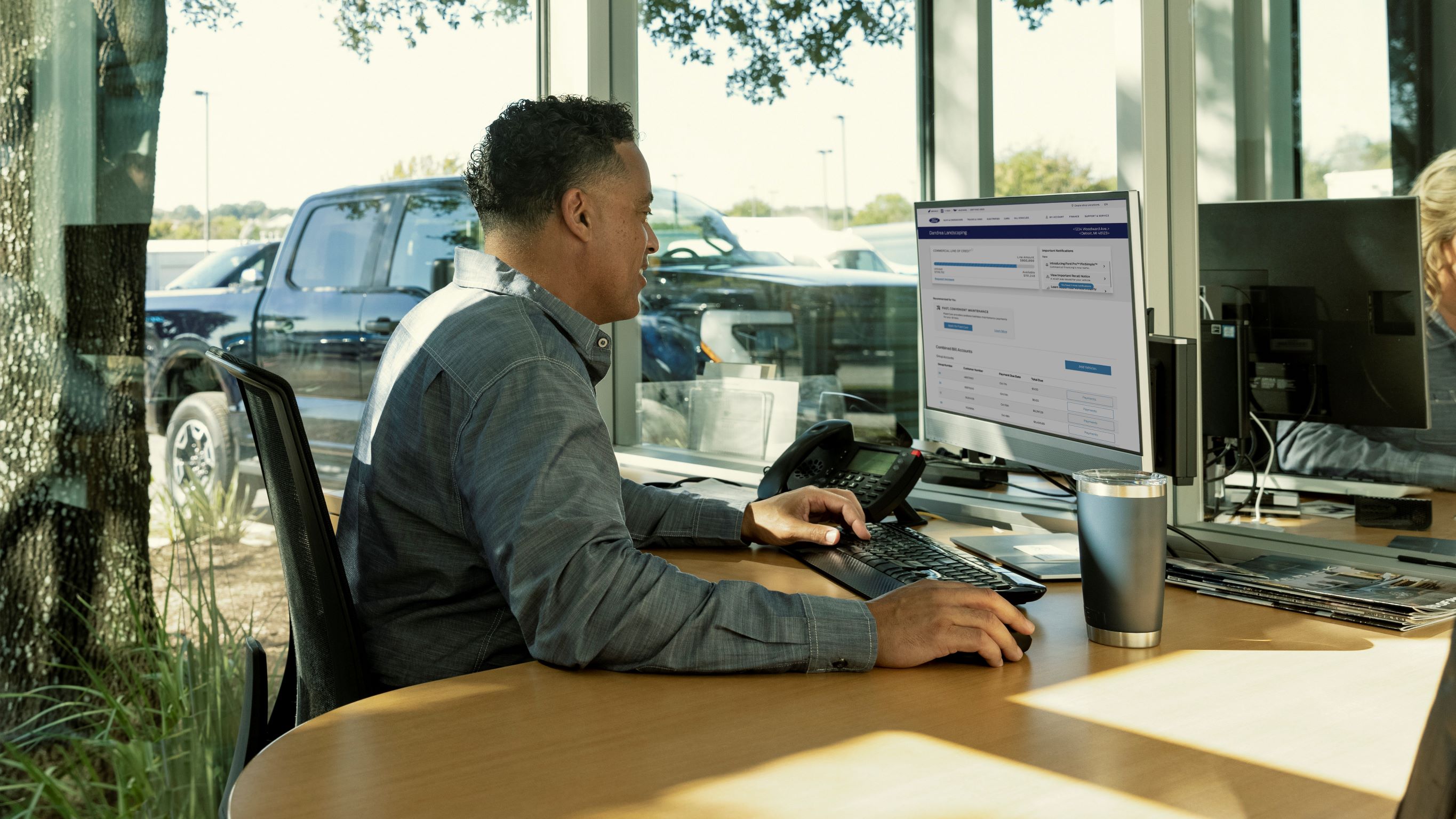 A man, presumably a fleet manager, sitting at a desk. He is viewing a page on large computer monitor. In the background, outside a window, a Ford Etransit is parked.