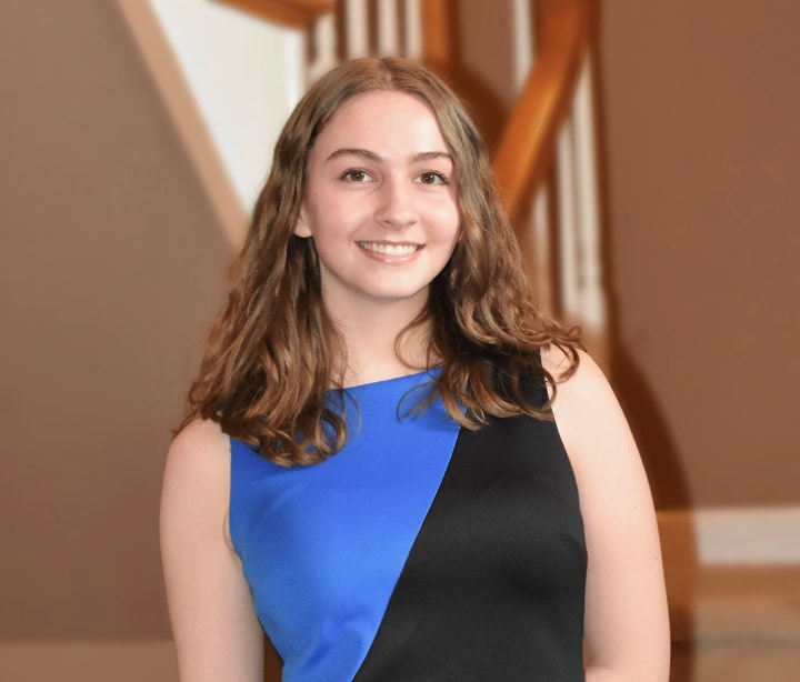 A woman with brown, wavy hair, wearing a blue and black dress, smiles in front of a wooden staircase.