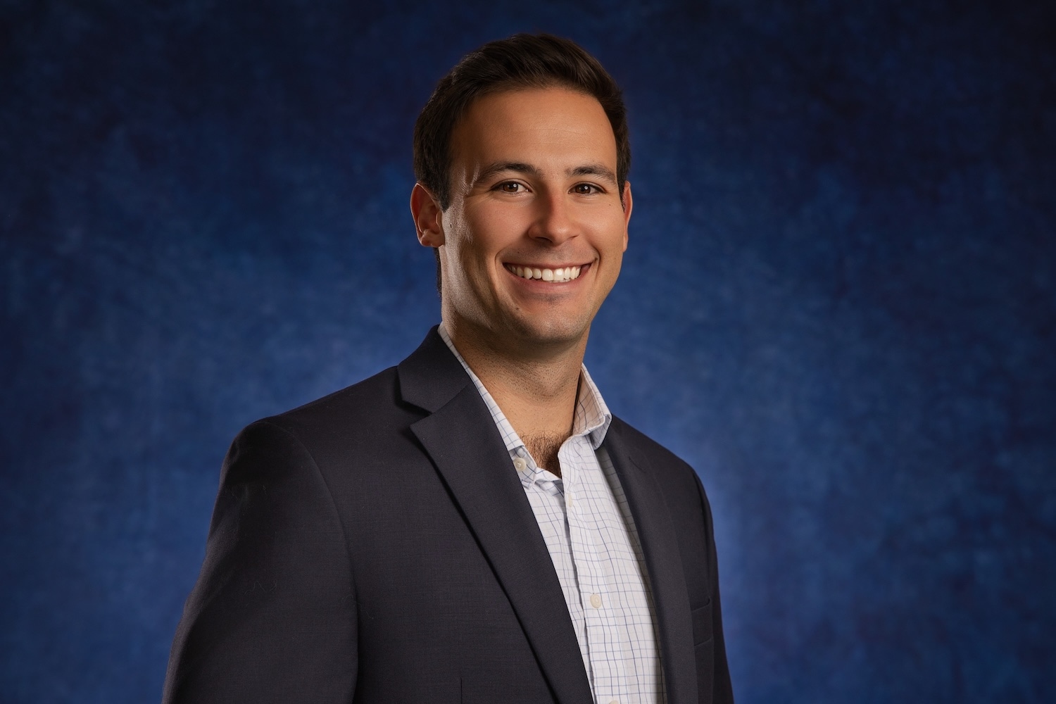 A man in a dark suit jacket and a light plaid shirt smiles against a blue background.