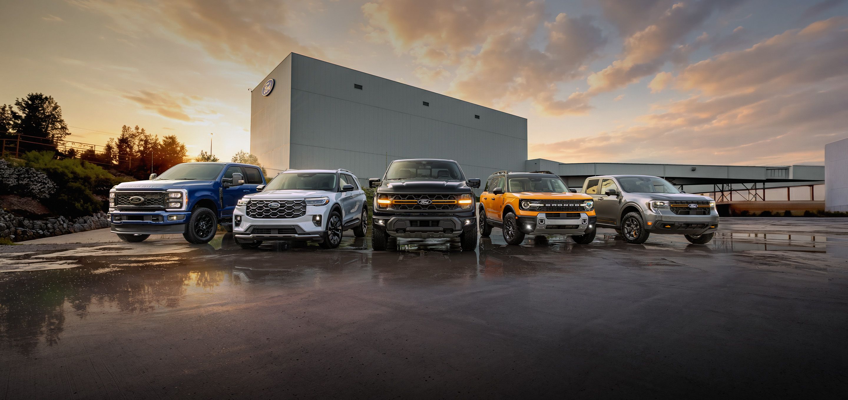 Une gamme de véhicules Ford est stationnée devant une usine Ford, sur un asphalte détrempé par la pluie, sous un ciel nuageux au coucher du soleil. Le texte indique : « Obtenez de belles offres sur les Ford neufs. »