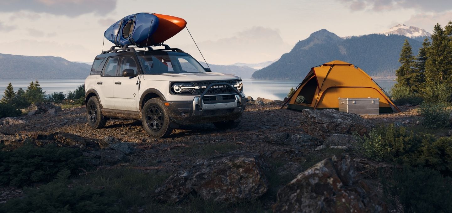 Ford Bronco Sport parked on rocky terrain beside a lake, with kayaks on the roof and a campsite set up nearby.