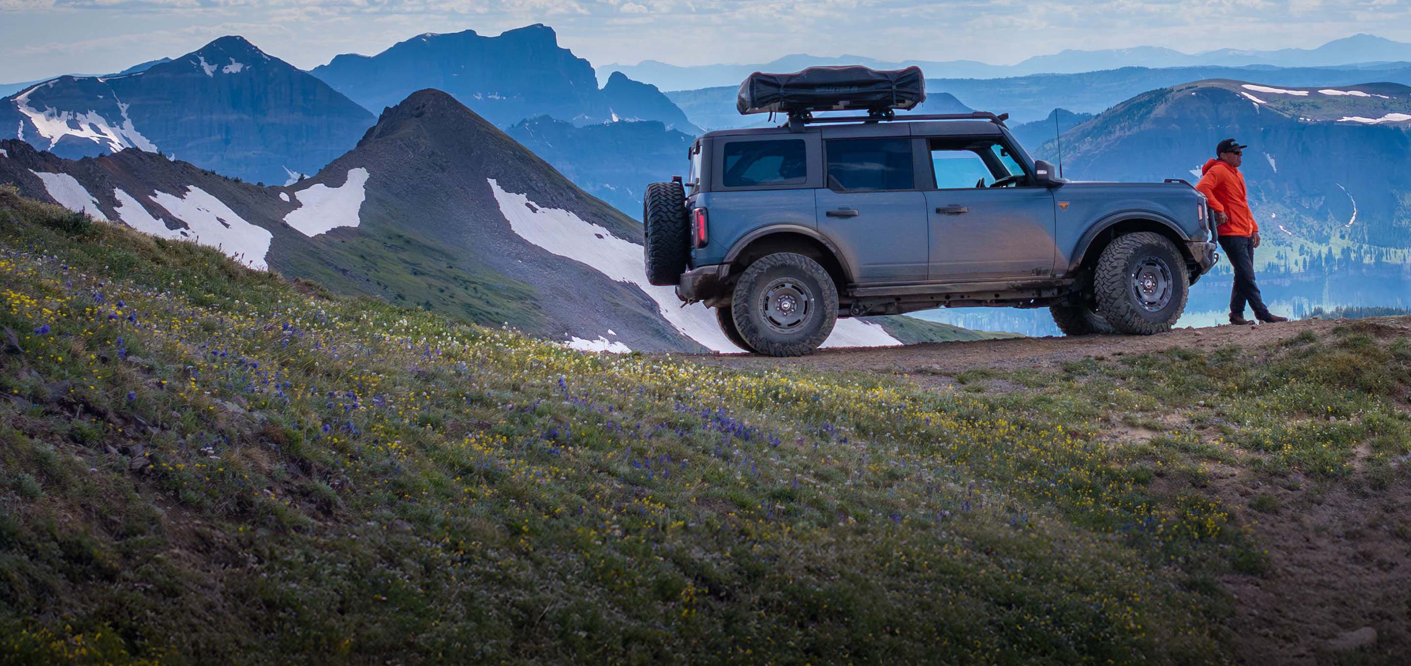 First Time Buyer Program. A person in an orange jacket stands beside their Ford Bronco on a mountain slope, gear loaded on the roof rack and ready for an adventure.