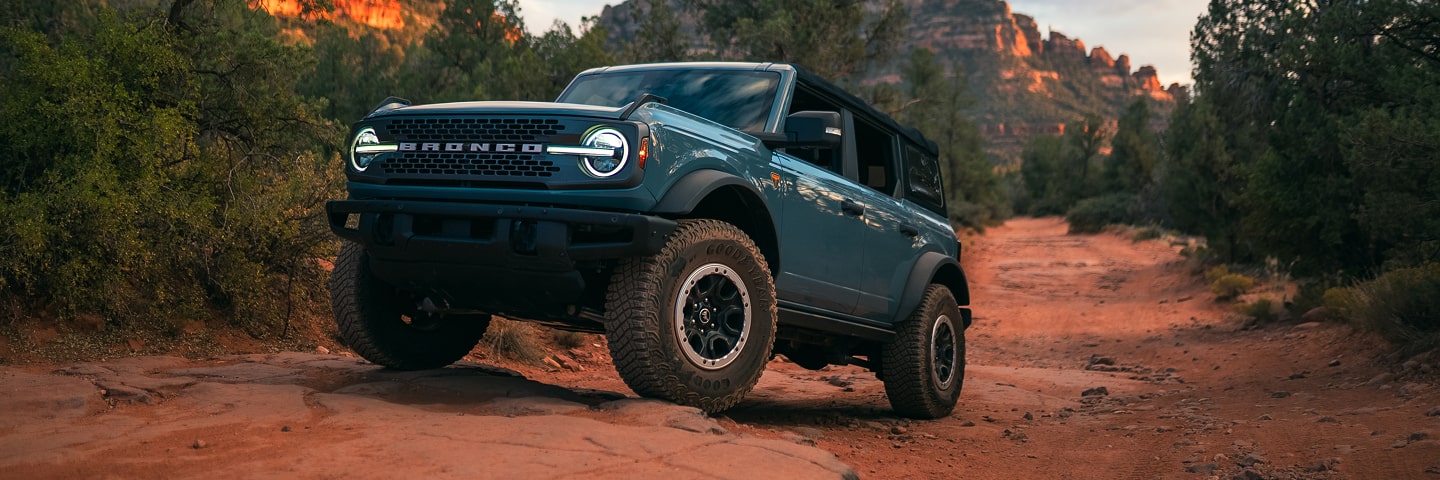 A blue Ford Bronco driving off-road on a dirt road with mountains in the background.