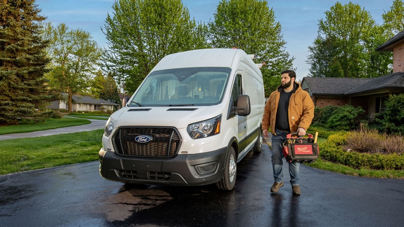Worker carrying a toolbox in front of a white Ford Transit cargo van.