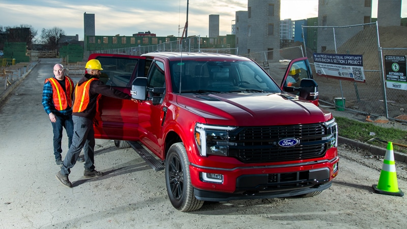 Workers in orange safety vests stand near a red Ford F-150 pickup truck at a construction site.