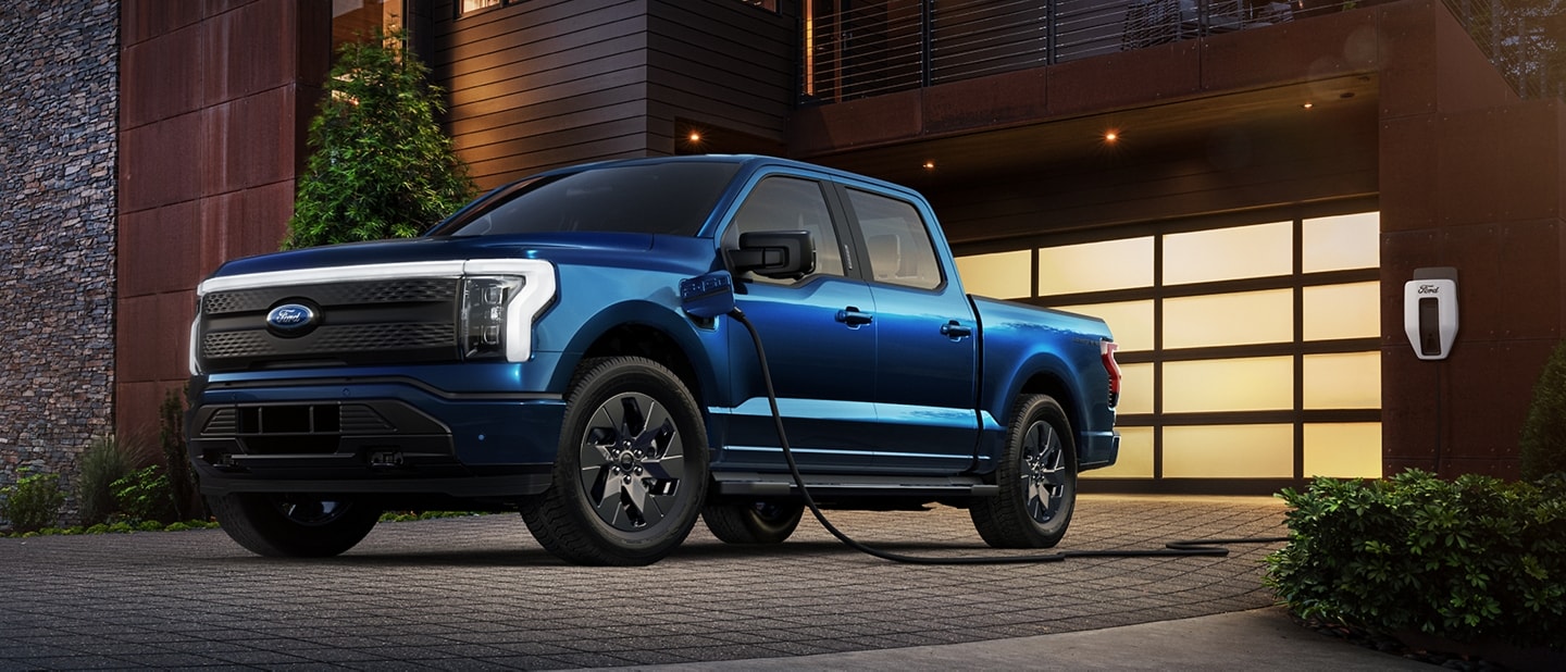 A blue 2023 Ford F-150® Lightning® parked in a driveway at night. It is plugged into a Ford Charge Station Pro