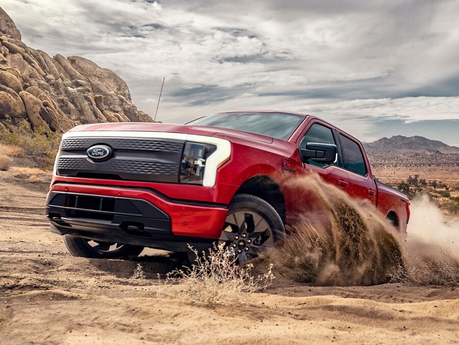 A red 2023 Ford F-150® Lightning® traversing a shallow river bed in a desert or prairie-like landscape