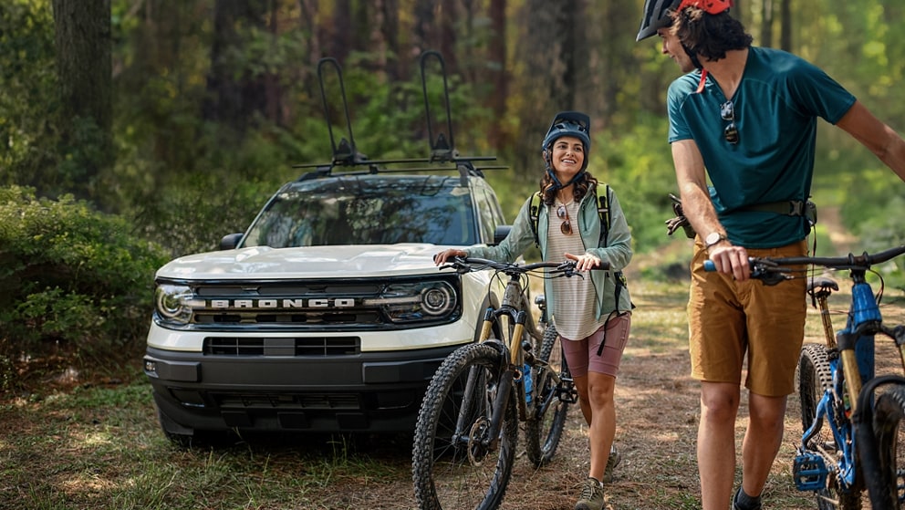 A Ford Bronco Sport is parked on a trail. A couple is walking their bikes away from it.