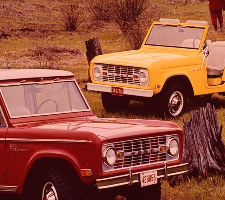 Three 1969 Ford Broncos parked in a wilderness field while a group of people walk nearby