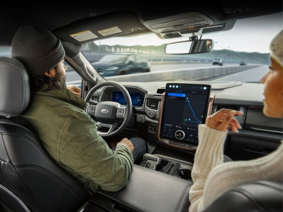 A delivery person stands beside the open door of a Ford vehicle while the new owner sits in the driver's seat