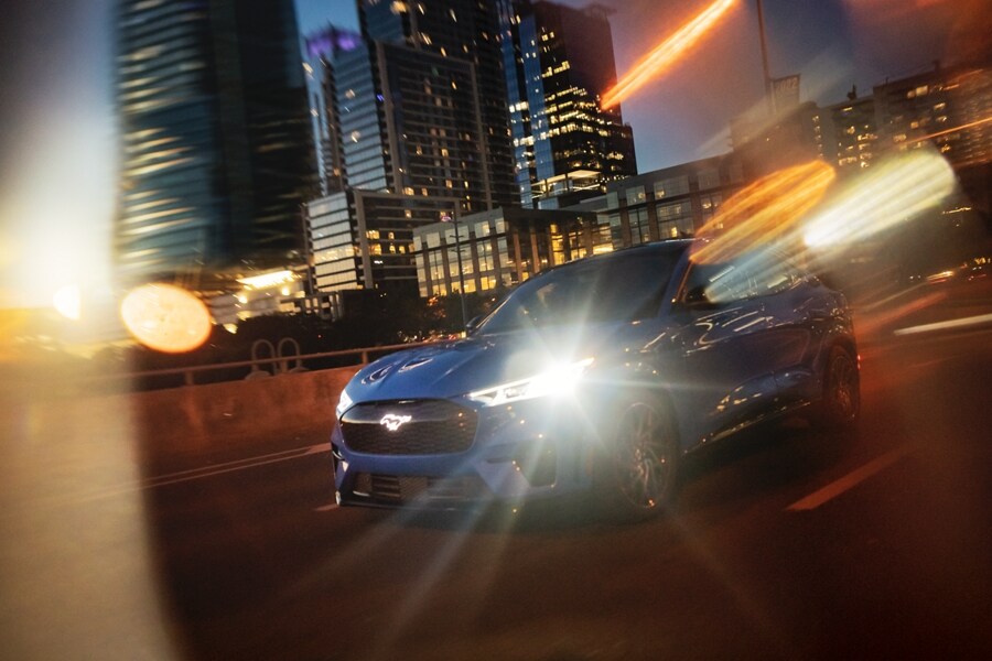 A Ford Mustang Mach-E drives on a highway past skyscrapers as light reflects off the vehicle’s headlights.  