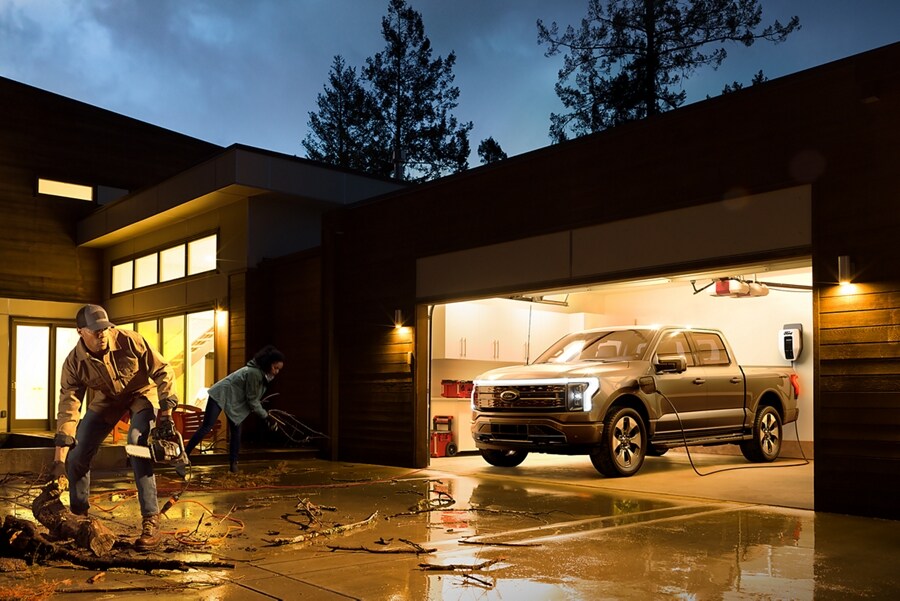 Couple cleaning up storm debris in a driveway with a Ford F-150® Lightning™ in the garage hooked up to charger