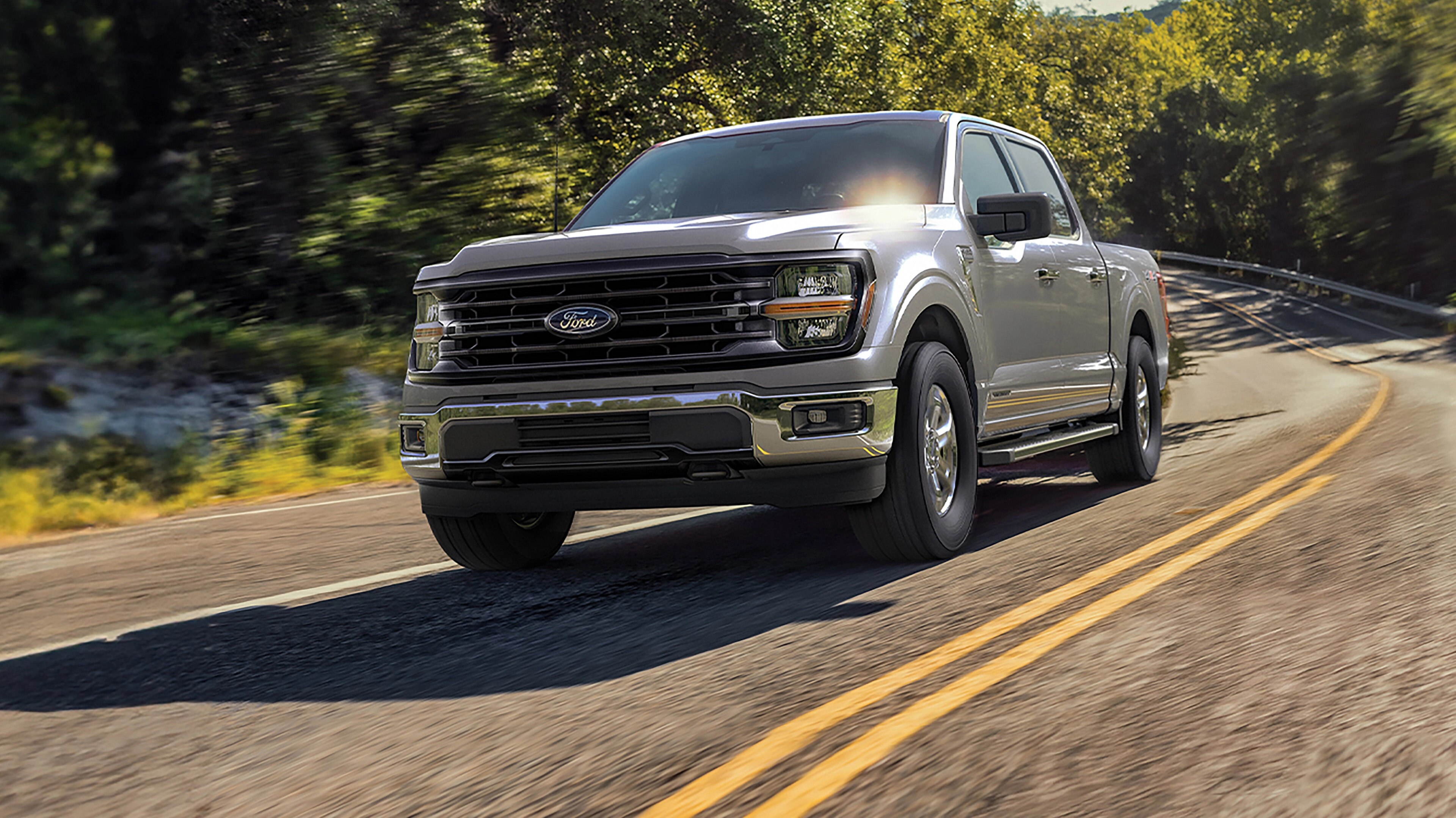 A gray 2025 Ford F-150® truck being driven down a road surrounded by trees