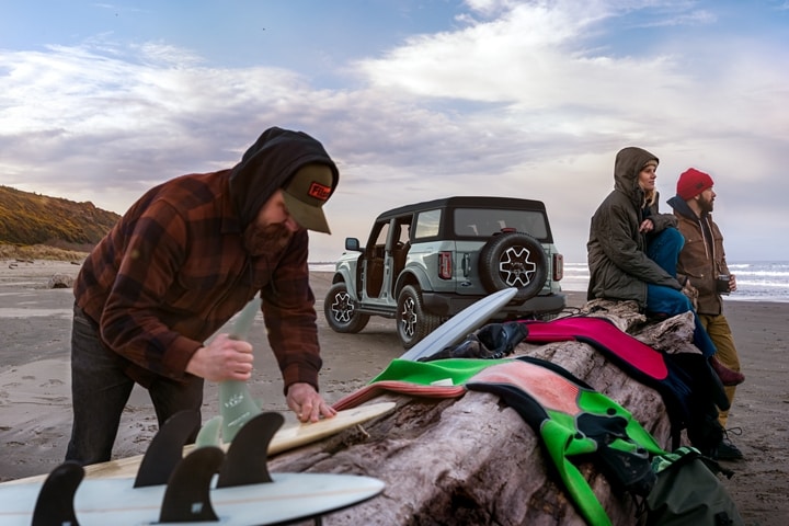Una Ford Bronco® Outer Banks® 2023 de cuatro puertas en Cactus Gray estacionada en la playa con tres surfistas
