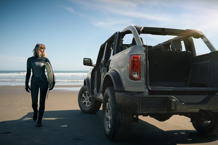 Una Ford Bronco® 2023 de cuatro puertas en Cactus Gray sin las puertas ni el techo disponible estacionada en la playa