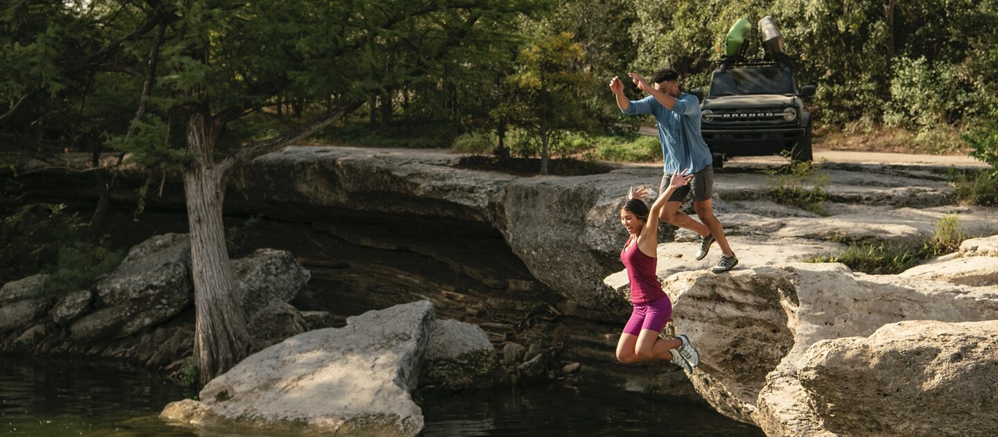Two people jumping into water with the a 2023 Ford Bronco® parked in the distance