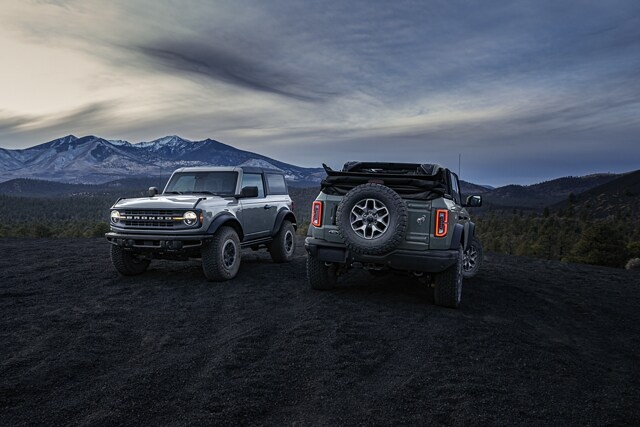Una Bronco® 2023 de dos puertas con techo duro y Black Diamond® de cuatro puertas con techo blando al atardecer en la montaña