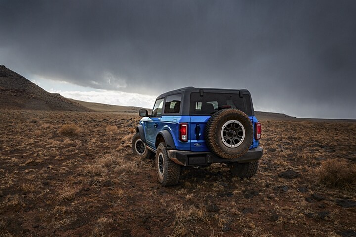 Vista trasera de una Ford Bronco® Black Diamond® 2023 de dos puertas en Velocity Blue andando por terreno escabroso con el Paquete Sasquatch® disponible