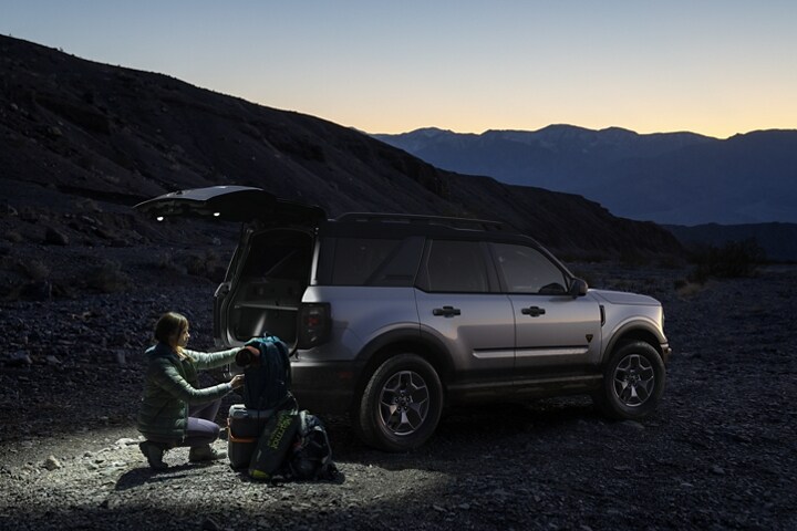 A person with camp gear near the open liftgate of a 2023 Ford Bronco® Sport SUV with rear floodlights illuminated