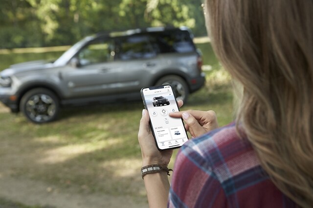 A woman holding a smartphone uses her FordPass® App to remote start her 2023 Ford Bronco® Sport SUV