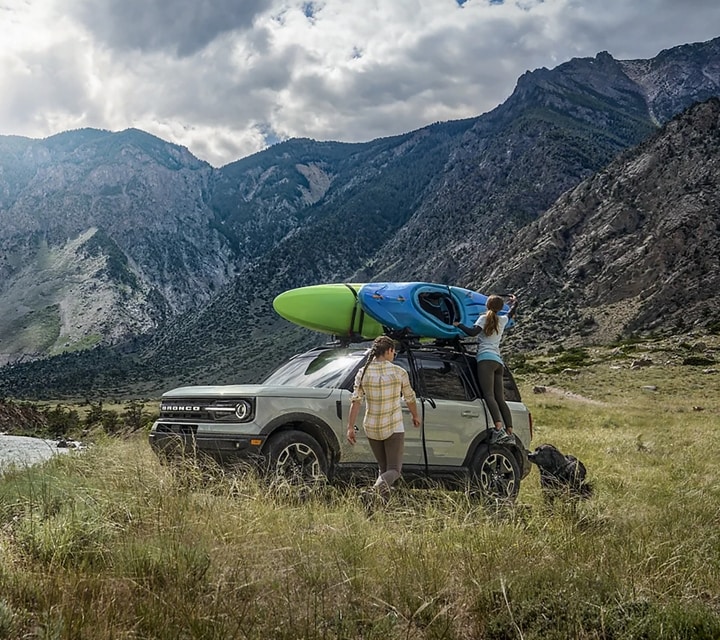 Two people are standing outside a 2024 Ford Bronco® Sport that is parked by a lake with a kayak on the roof