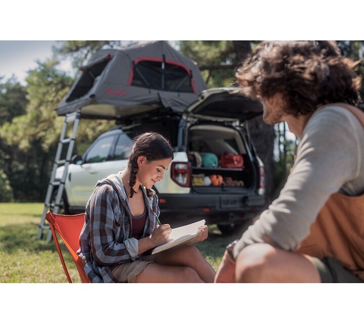 A 2024 Ford Bronco® Sport is in the background while a young woman writes in her journal and a man sits near her