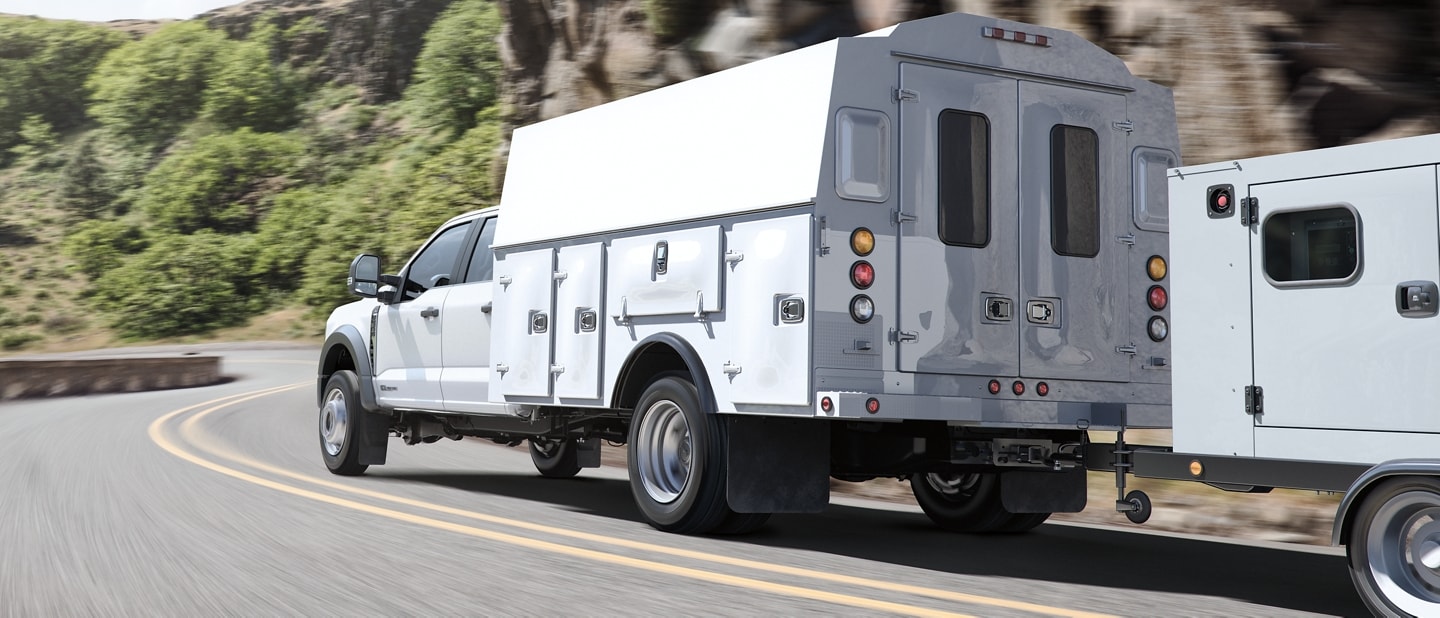 2023 Ford Super Duty® Chassis Cab with utility box upfit and trailer in tow being driven on road near trees