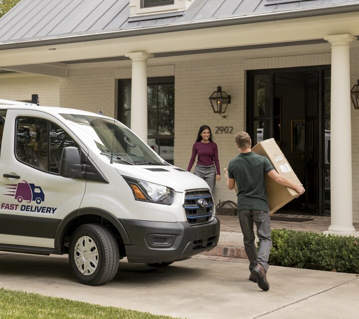 A white 2024 Ford E-Transit™ van parked at a home as a delivery worker delivers a package