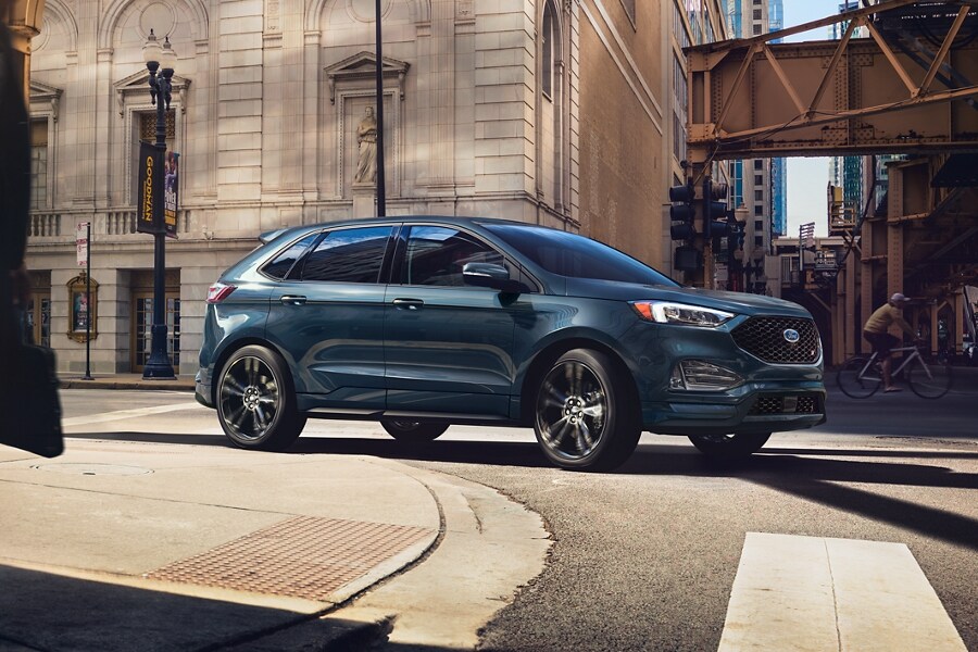A 2023 Ford Edge® SUV in Stone Blue being driven around the curve in a city street