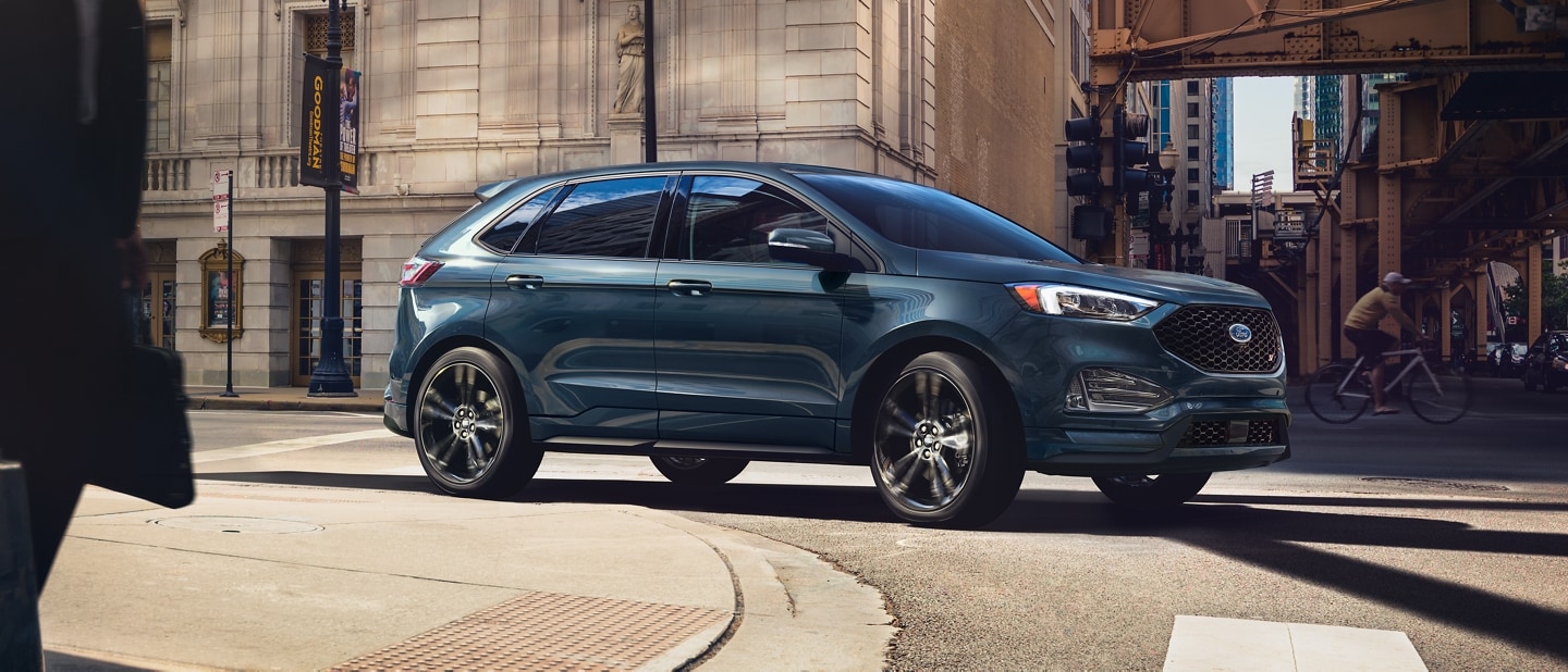 A 2024 Ford Edge® SUV in Stone Blue being driven around the curve in a city street