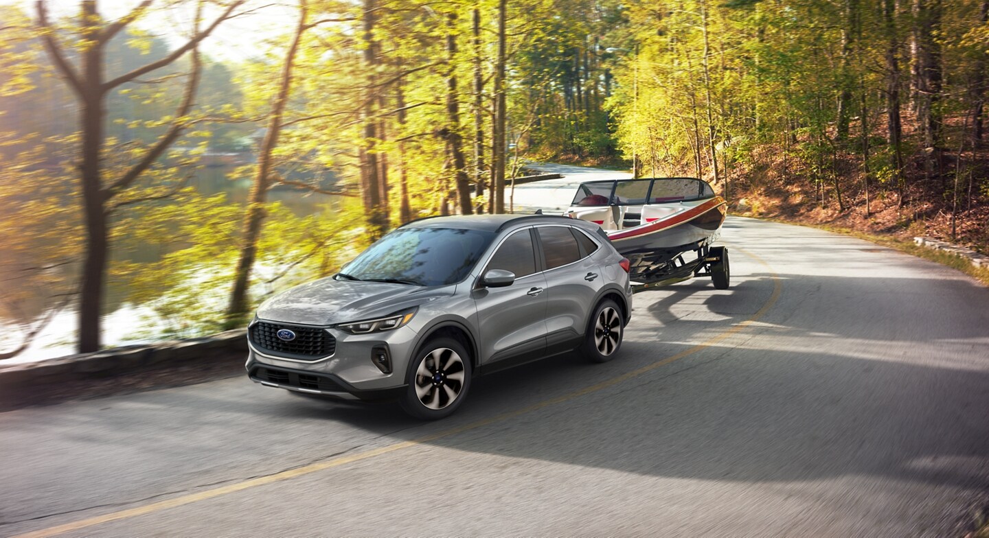 An iconic silver 2024 Ford Escape pulling a small watercraft along a winding wooded road