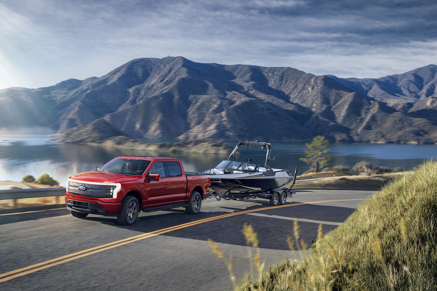  2024 red Ford Lightning Lariat towing a boat on highway with lake and mountains in the background. 