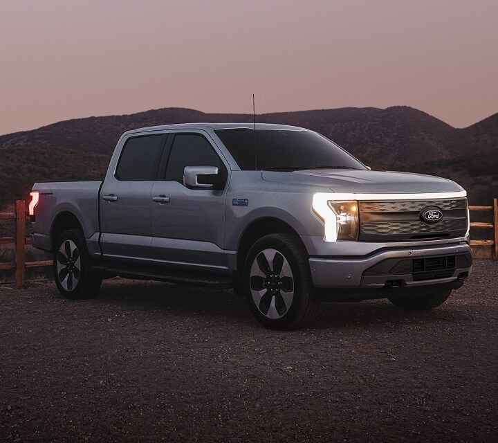 A blue-gray 2024 Ford F-150 Lightning® pickup truck parked on a gravel road near a fenced-in field