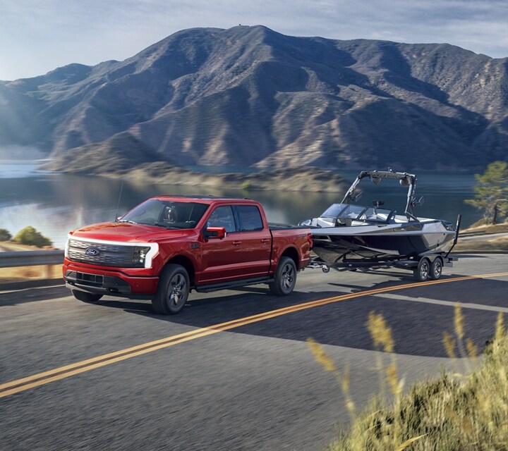 A 2024 Ford F-150 Lightning® pickup truck pulling a motorboat down a rural highway, with a mountain lake in the background