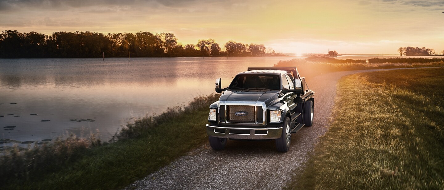 Una Ford F-750 Crew Cab 2024 en Agate Black con mejora de plataforma con estacas utilitarias andando cerca de un lago