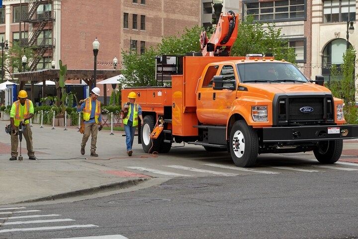 Unos trabajadores caminando cerca de una Ford F-750 Crew Cab 2024 con mejora de plataforma elevadora en Tangier Orange estacionada en una calle de ciudad