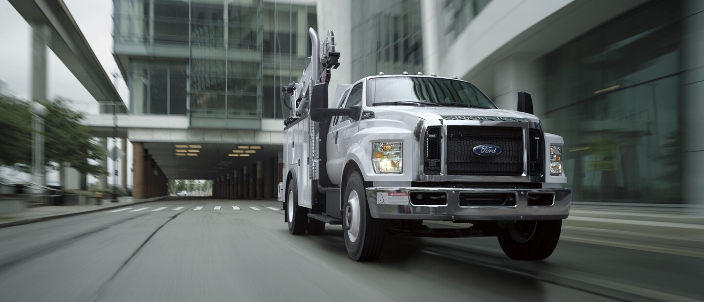 Una Ford F-750 SuperCab 2024 en Oxford White con con mejora mecánica circulando junto a un edificio grande