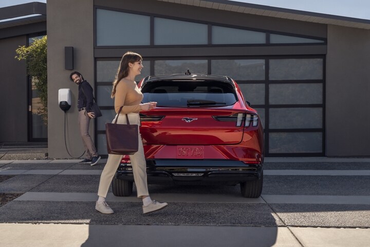 Mujer que pasa caminando por la parte trasera de una Ford Mustang Mach-E® 2023 estacionada en la entrada de una casa mientras un hombre desenchufa el vehículo