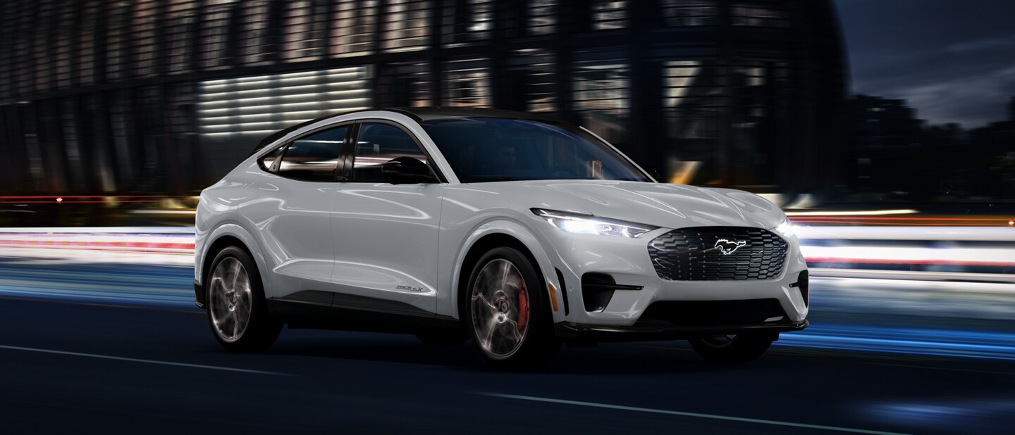 Una Ford Mustang Mach-E® 2023 en Space White Metallic circulando por una carretera pasando por un estadio de noche