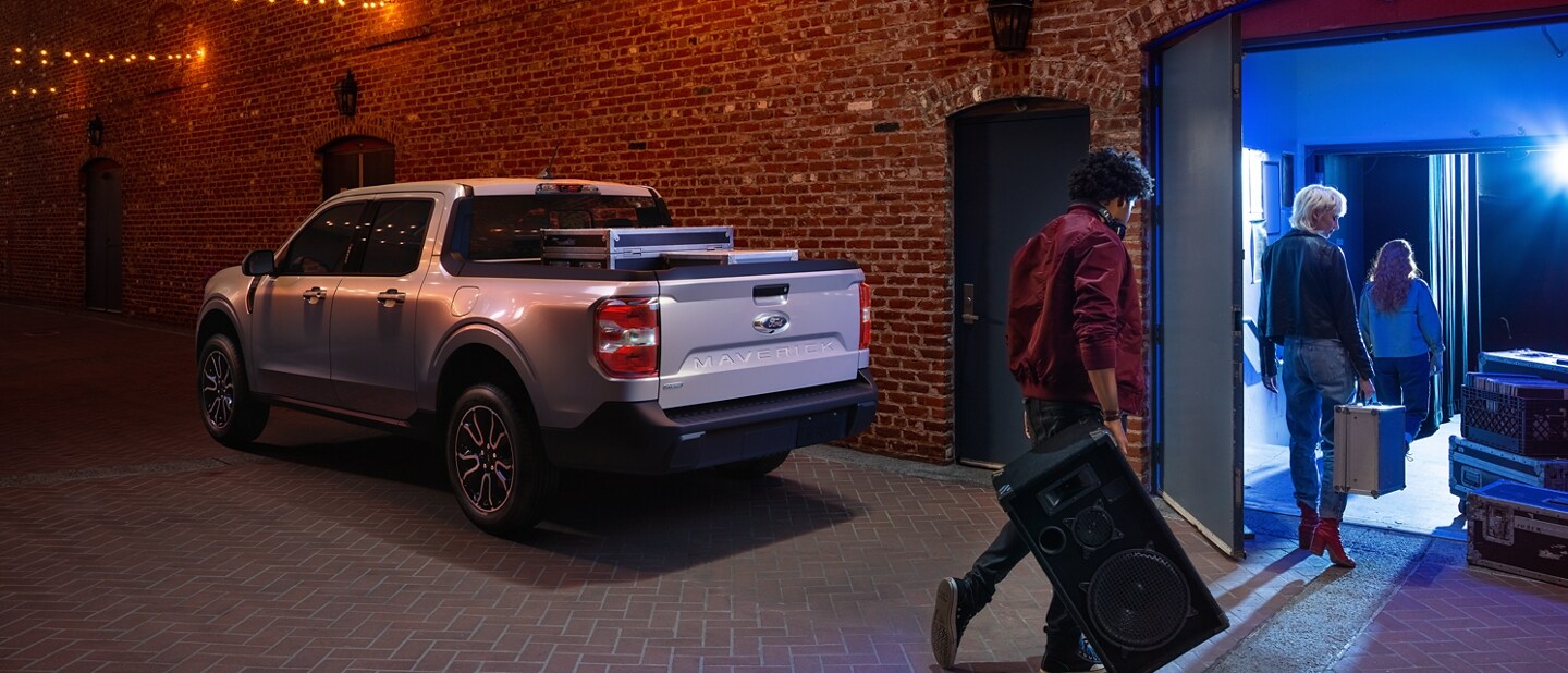 2023 Ford Maverick® truck in Iconic Silver parked in an alley with musicians bringing gear into a nightclub